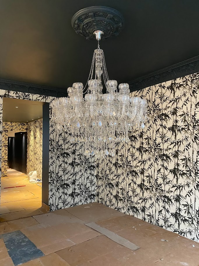 Kryształowy żyrandol w pokoju z czarnym sufitem i tapetą w motywy bambusowe, podłoga zabezpieczona kartonem podczas prac wykończeniowych.
