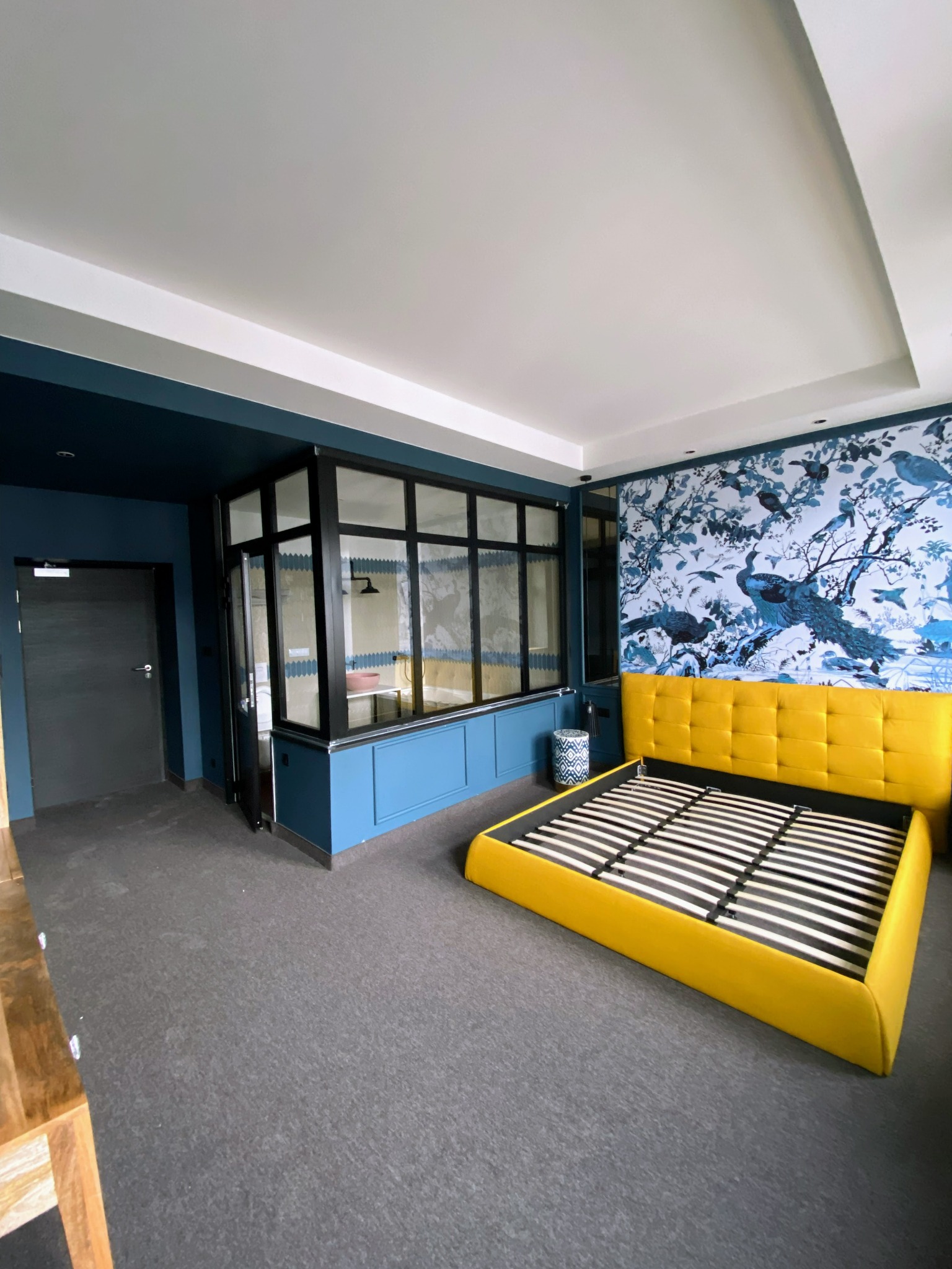 Pokój z niebieskimi ścianami, dekoracyjną tapetą z motywem ptaków i żółtym łóżkiem bez materaca, oddzielony przeszkloną ścianą od łazienki.
