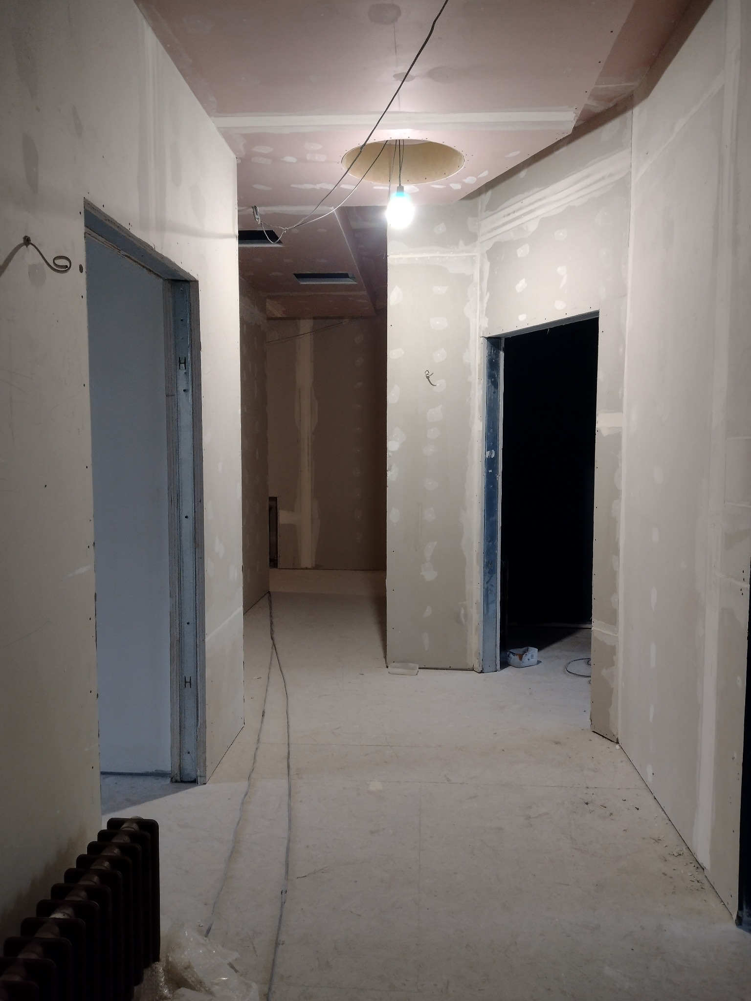 Korytarz w trakcie prac wykończeniowych: ściany pokryte płytami kartonowo-gipsowymi, widoczne łączenia do szpachlowania, sufit z okrągłym otworem na oświetlenie, grzejnik żeliwny w rogu.