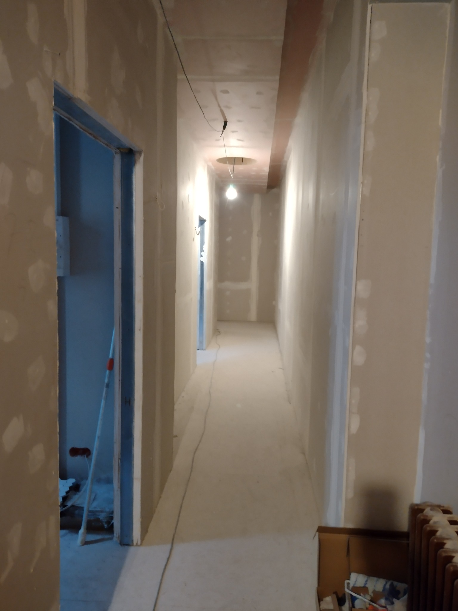 Korytarz w trakcie prac wykończeniowych, ściany pokryte gładzią z widocznymi łatami, sufit z elementem dekoracyjnym w kolorze terakoty, widoczne kable oświetleniowe, w tle otwarte drzwi...
