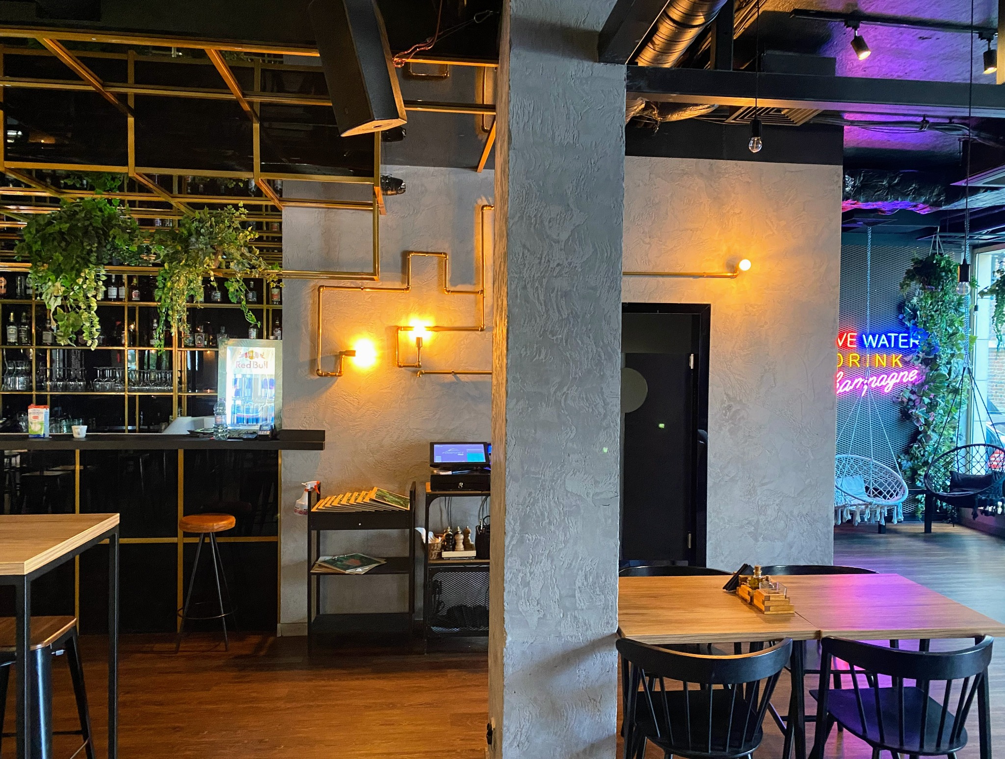Wnętrze lokalu gastronomicznego z dekoracyjną kolumną o szarej, chropowatej fakturze, złotymi elementami instalacji na ścianie, neonem i roślinami w tle.