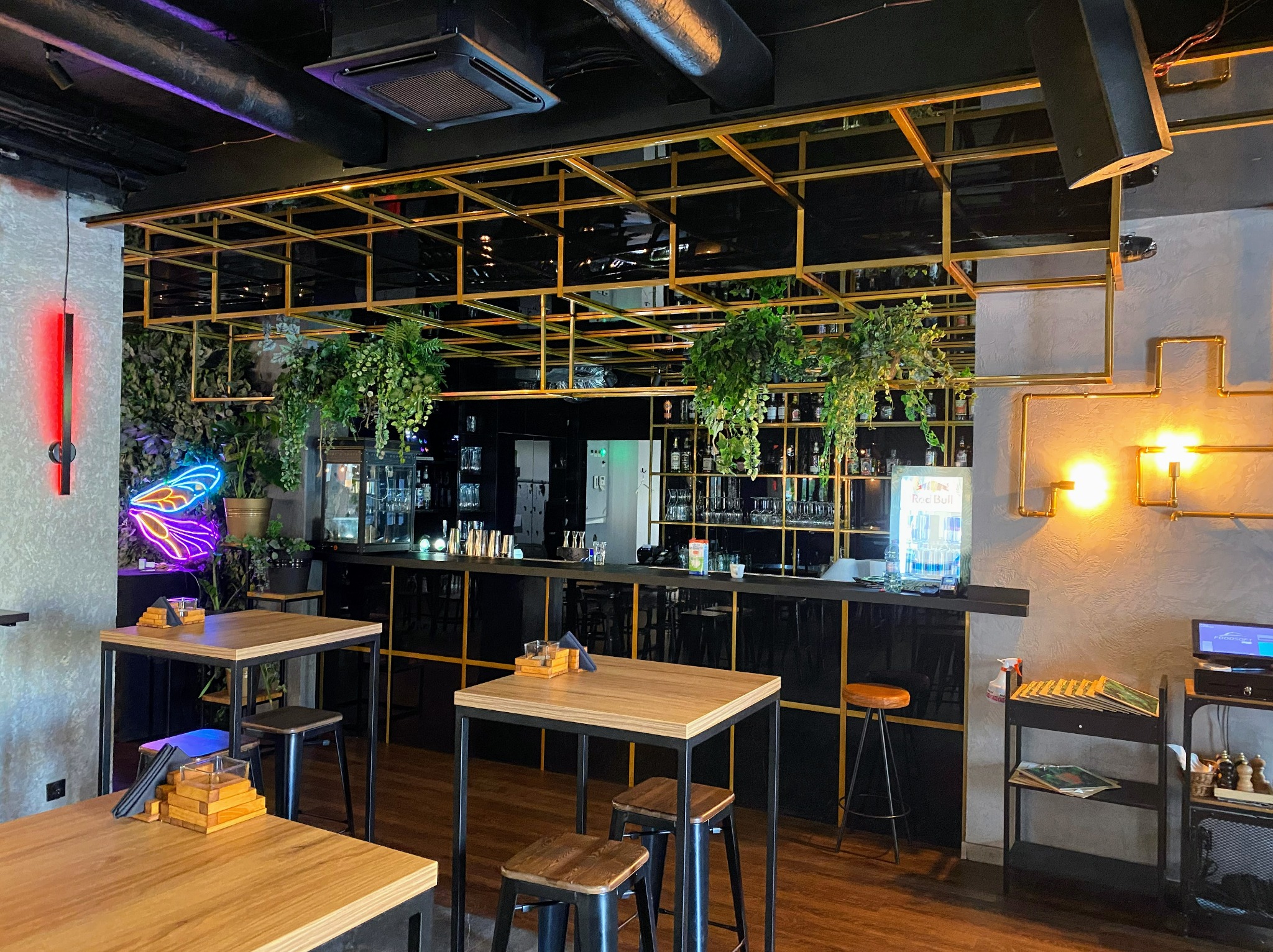 Wnętrze baru z drewnianymi stolikami i krzesłami, czarnym barem z złotymi akcentami, roślinami zwisającymi z sufitu i neonem w kształcie motyla na ścianie.