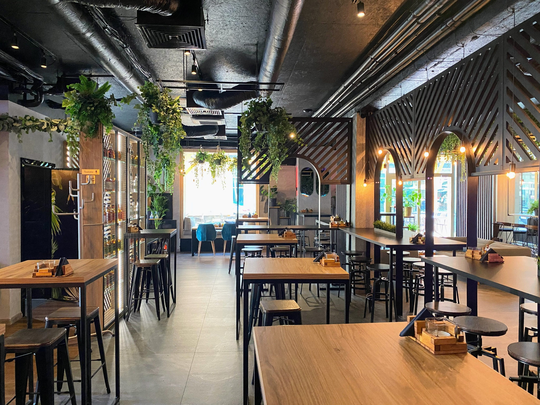 Wnętrze nowoczesnej restauracji z drewnianymi stolikami i czarnymi krzesłami barowymi, dekoracjami roślinnymi i industrialnym oświetleniem sufitowym.