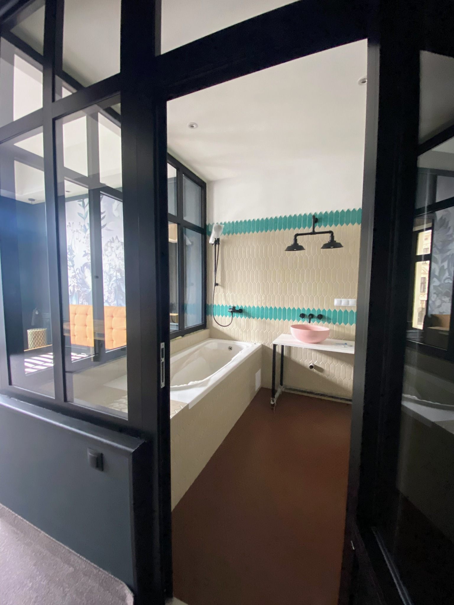 Nowoczesna łazienka z czarnymi ramami okiennymi i szklanymi drzwiami, beżowymi płytkami w kształcie łezki z turkusowymi akcentami, wanną i różową umywalką na białym blacie z czarnymi nogami, widok...