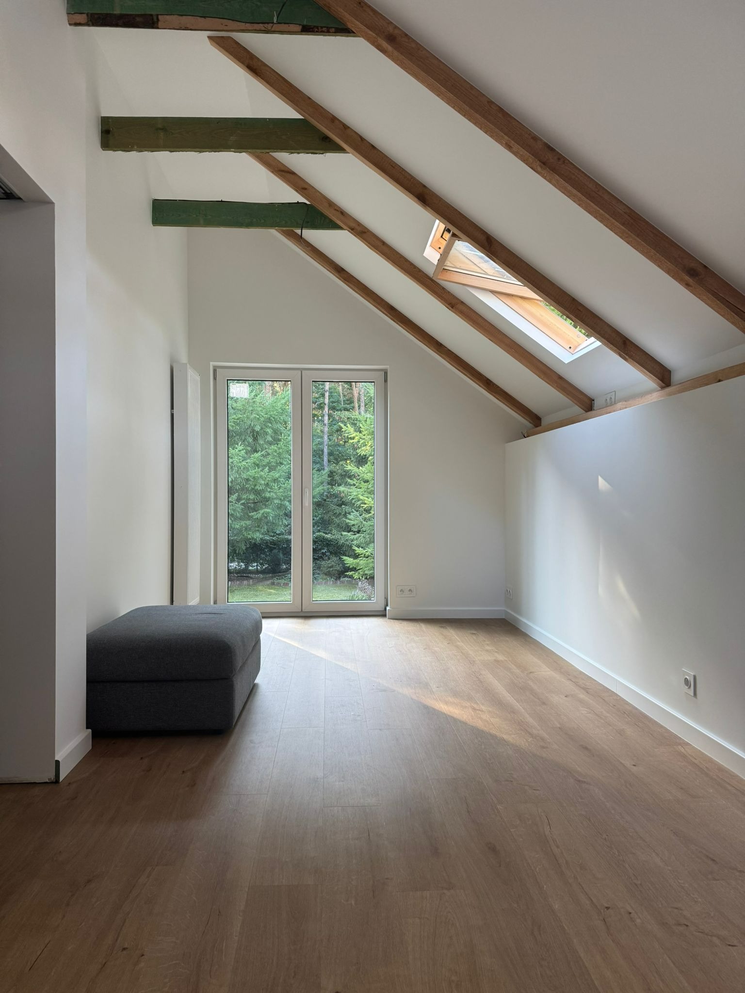 Jasne poddasze po remoncie: drewniane belki, okno dachowe, panele podłogowe, białe ściany. Widok na zieleń przez okno balkonowe. Szara pufa w rogu.