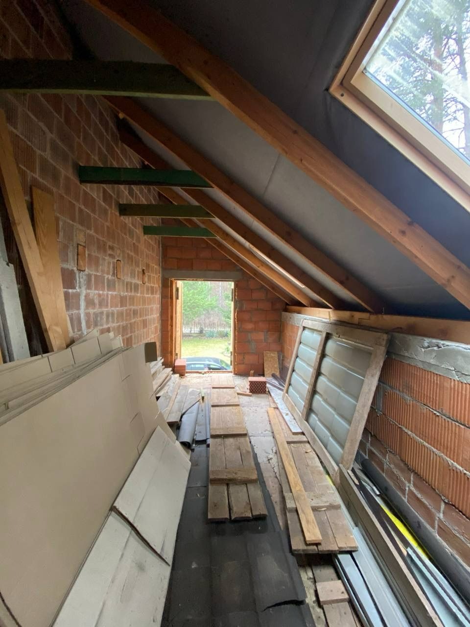 Poddasze w trakcie adaptacji: surowe ściany z cegły, drewniane belki stropowe, okno dachowe i materiały budowlane przygotowane do dalszych prac wykończeniowych.