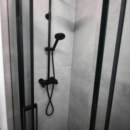 BUDMARK Marek Mróz Firma Remontowo-Budowlana - Czarna armatura prysznicowa, w tym deszczownica i słuchawka, na tle szarych płytek w kabinie z czarnymi profilami.