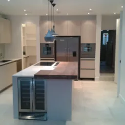 Nowoczesna kuchnia z wyspą, zabudową meblową w jasnych odcieniach i drewnianym blatem. Widoczne oświetlenie punktowe i wiszące lampy. Dominuje minimalizm i funkcjonalność.