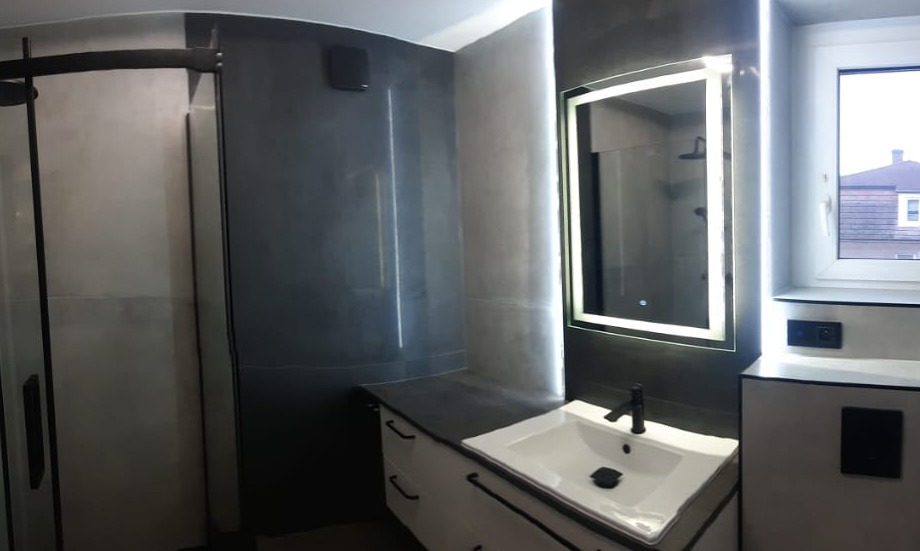 Nowoczesna łazienka z kabiną prysznicową typu walk-in, szarą glazurą, podświetlanym lustrem i białą szafką z czarnymi uchwytami.
