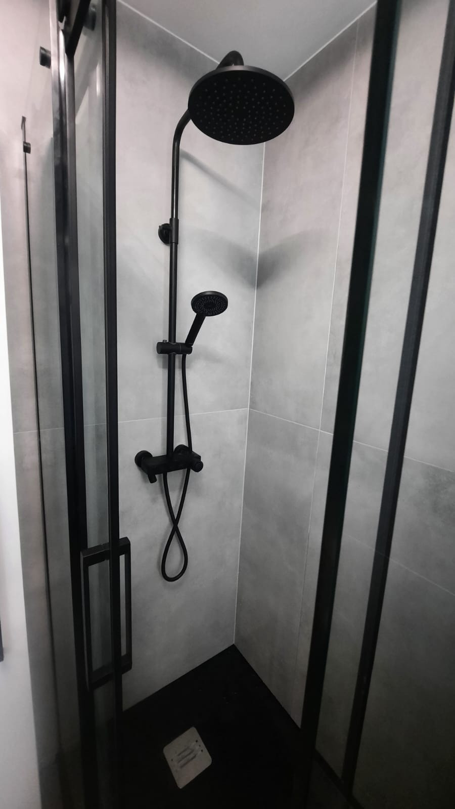Czarna armatura prysznicowa, w tym deszczownica i słuchawka, na tle szarych płytek w kabinie z czarnymi profilami.