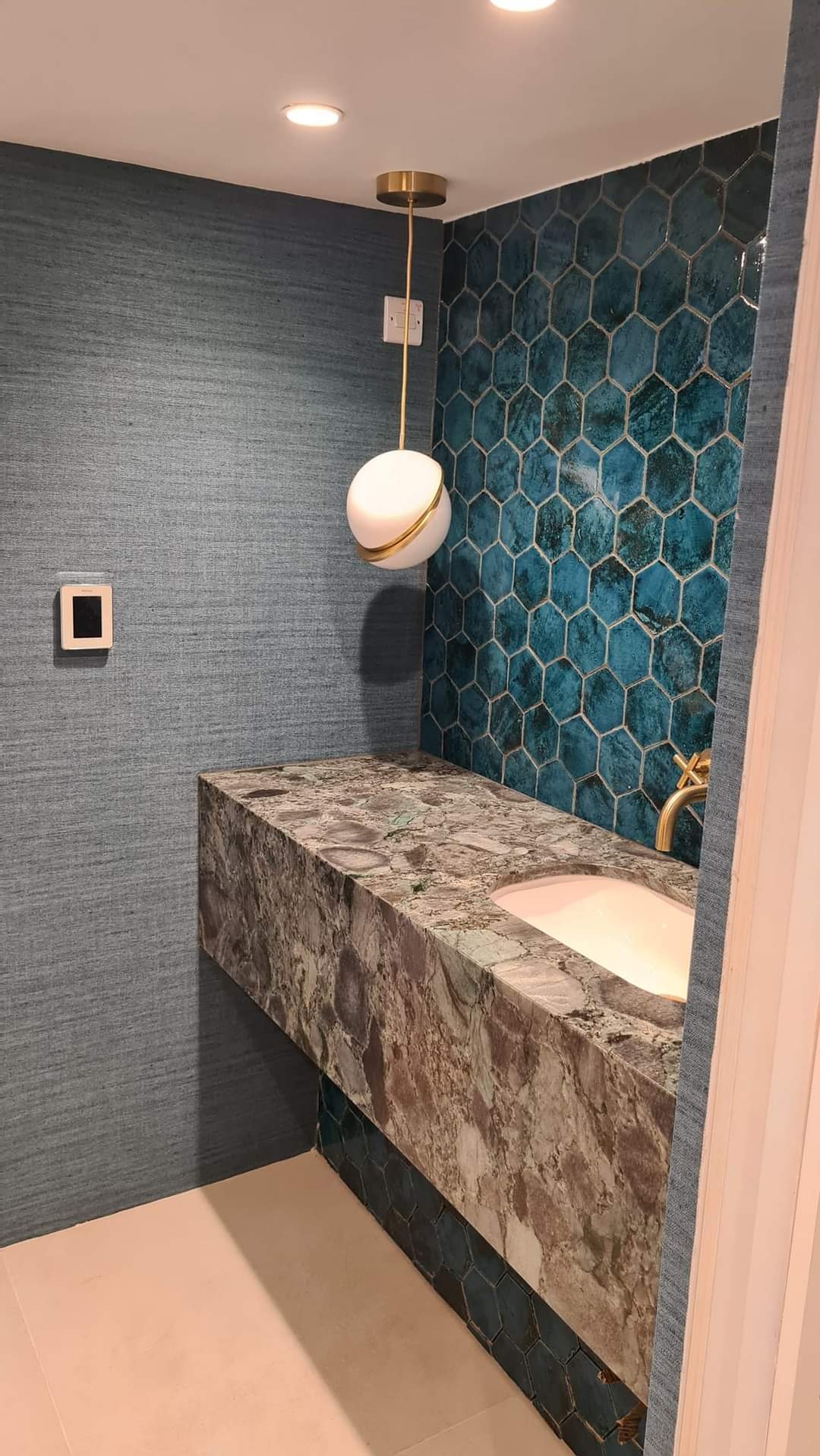 Nowoczesna łazienka z umywalką nablatową z kamienia, mozaiką heksagonalną w odcieniach turkusu, lampą wiszącą w kształcie kuli i ścianą wykończoną tapetą o fakturze lnu.