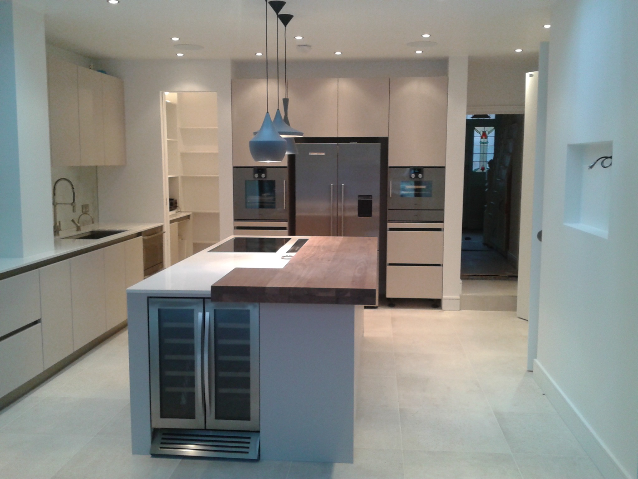 Nowoczesna kuchnia z wyspą, zabudową meblową w jasnych odcieniach i drewnianym blatem. Widoczne oświetlenie punktowe i wiszące lampy. Dominuje minimalizm i funkcjonalność.