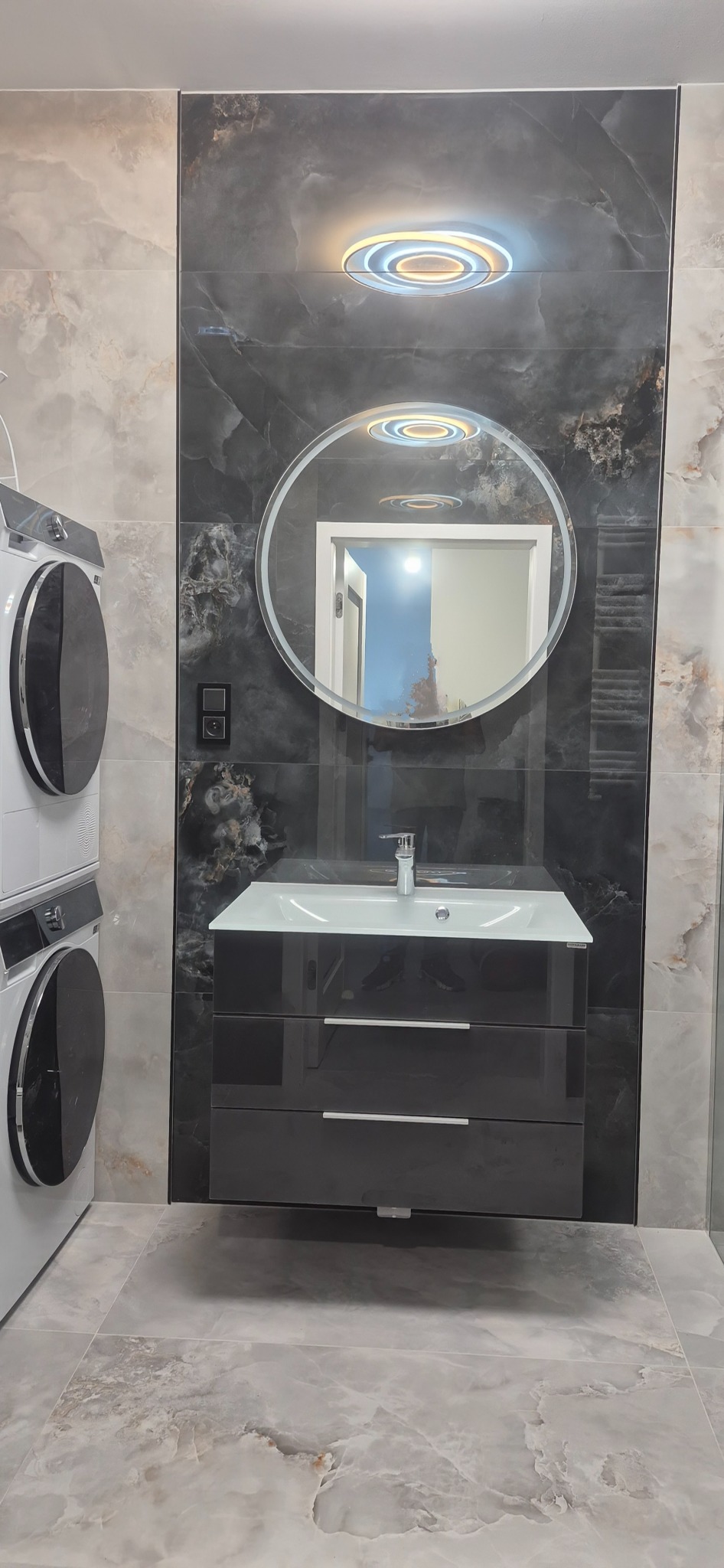 Nowoczesna łazienka z umywalką w szafce, okrągłym lustrem z podświetleniem LED i płytkami imitującymi marmur. Widoczna pralka i suszarka w kolumnie.