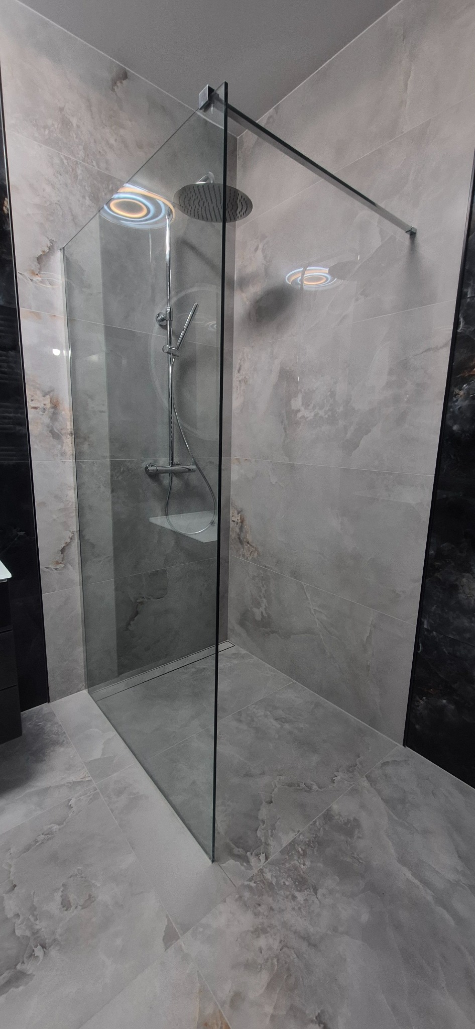 Nowoczesna kabina prysznicowa typu walk-in z deszczownicą, wykończona płytkami imitującymi marmur w odcieniach szarości i bieli, z widocznym odpływem liniowym.
