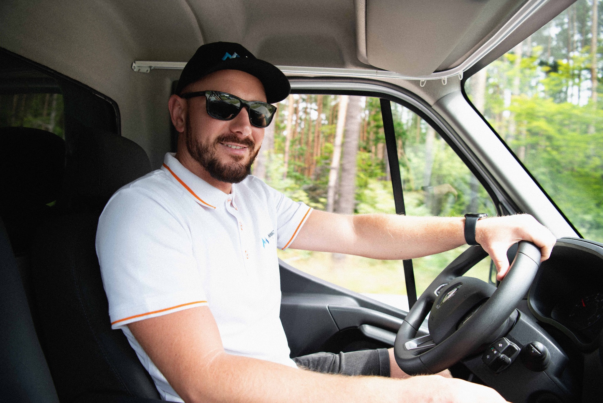 Kierowca w okularach przeciwsłonecznych i koszulce polo z logo firmy MAKO TSL siedzi za kierownicą samochodu dostawczego, widok na las przez okno.