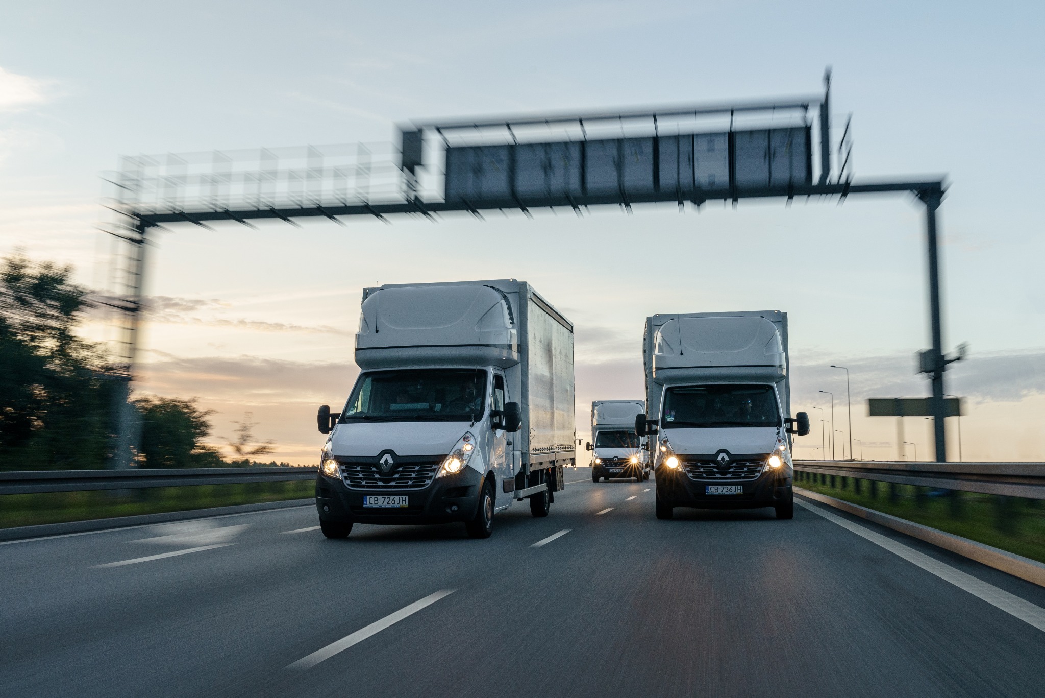 Kolumna trzech białych samochodów dostawczych Renault Master z wysoką zabudową kontenerową, jadących po autostradzie o zmierzchu, widok z przodu.