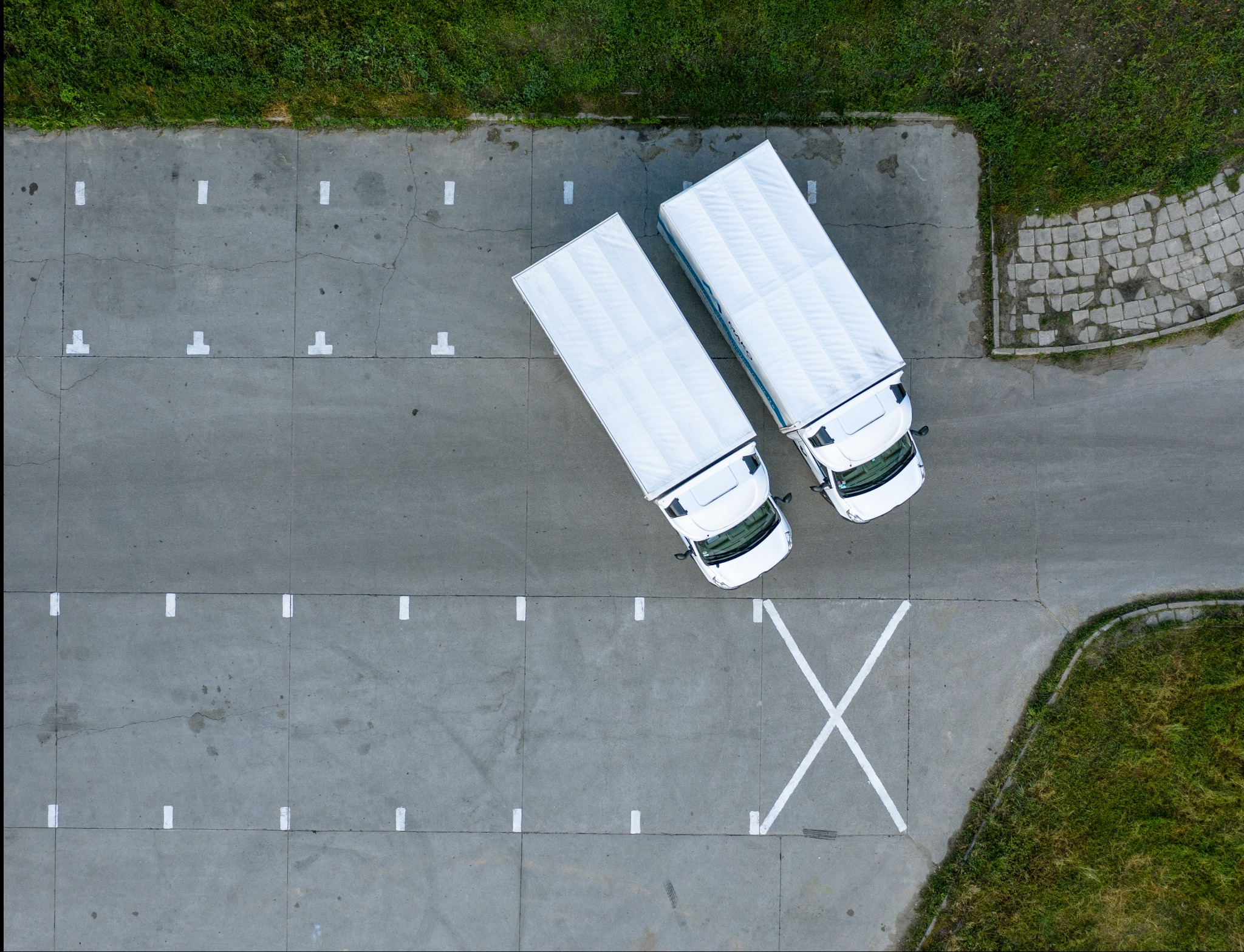 Dwa białe samochody ciężarowe z naczepami na betonowym placu parkingowym, widok z góry, z widocznymi liniami wyznaczającymi miejsca postojowe oraz zielenią na obrzeżach.