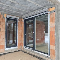 Grupa Sain - Hespol - Montaż okien PCV z roletami zewnętrznymi w surowym budynku. Widoczne bloczki betonowe stropu i cegły. Okna z czarnymi ramami i białą listwą montażową.