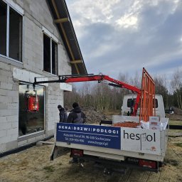 Grupa Sain - Hespol - Montaż okna w surowym budynku za pomocą przyssawki i dźwigu, widoczni pracownicy, auto dostawcze z reklamą okien, drzwi i podłóg w tle.