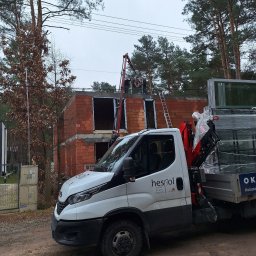 Grupa Sain - Hespol - Montaż okien w surowym budynku z użyciem dźwigu. Na pierwszym planie samochód dostawczy firmy z oknami. Widok na plac budowy w pochmurny dzień.