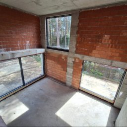 Grupa Sain - Hespol - Surowy stan: wnętrze z trzema oknami w czarnych ramach, widok na zieleń. Ściany z czerwonej cegły, surowy betonowy strop, wylewka na podłodze. Widok z góry.
