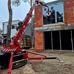 Grupa Sain - Hespol - Montaż okna na budowie domu jednorodzinnego za pomocą specjalistycznego dźwigu z przyssawkami. Widoczny surowy stan budynku i otoczenie placu budowy.