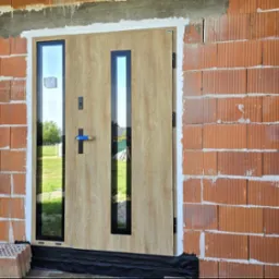 Jasne drzwi zewnętrzne z przeszkleniami, czarnymi ramami i klamką, zamontowane w surowym murze z cegły ceramicznej. Widoczne detale montażu.