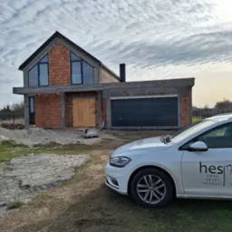 Surowy stan domu z cegły z zamontowanymi oknami, garaż z bramą i auto firmy Hespol na pierwszym planie, pochmurne niebo w tle.