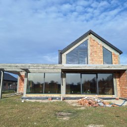 Grupa Sain - Hespol - Surowy stan domu z zamontowanymi oknami aluminiowymi, ceglana elewacja w trakcie budowy, widoczne okna z ciemnymi ramami i fragment dachu z szarą dachówką.