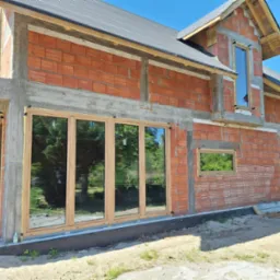 Surowy stan budynku z zamontowanymi oknami o drewnianych ramach. Widoczne cegły i betonowe elementy konstrukcyjne. Odbicie zieleni w szybach.