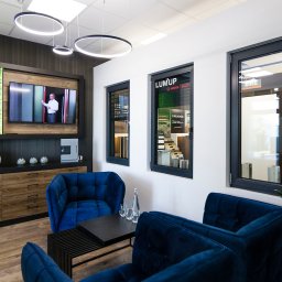 Grupa Sain - Hespol - Nowoczesne okna aluminiowe w salonie ekspozycyjnym. Czarne ramy, minimalistyczny design, komfortowa przestrzeń dla klienta z niebieskimi fotelami.