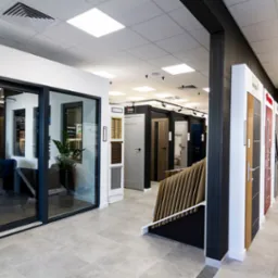 Salon ekspozycyjny z różnymi modelami drzwi wewnętrznych i przesuwnych, oświetleniem punktowym i szarą podłogą. Widoczna zieleń roślin w donicach.