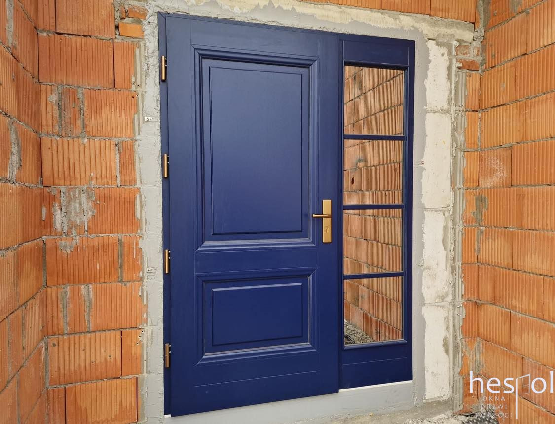 Granatowe drzwi zewnętrzne z szybą boczną, osadzone w surowym murze z cegły. Złoty uchwyt i zawiasy kontrastują z głębokim odcieniem drzwi.