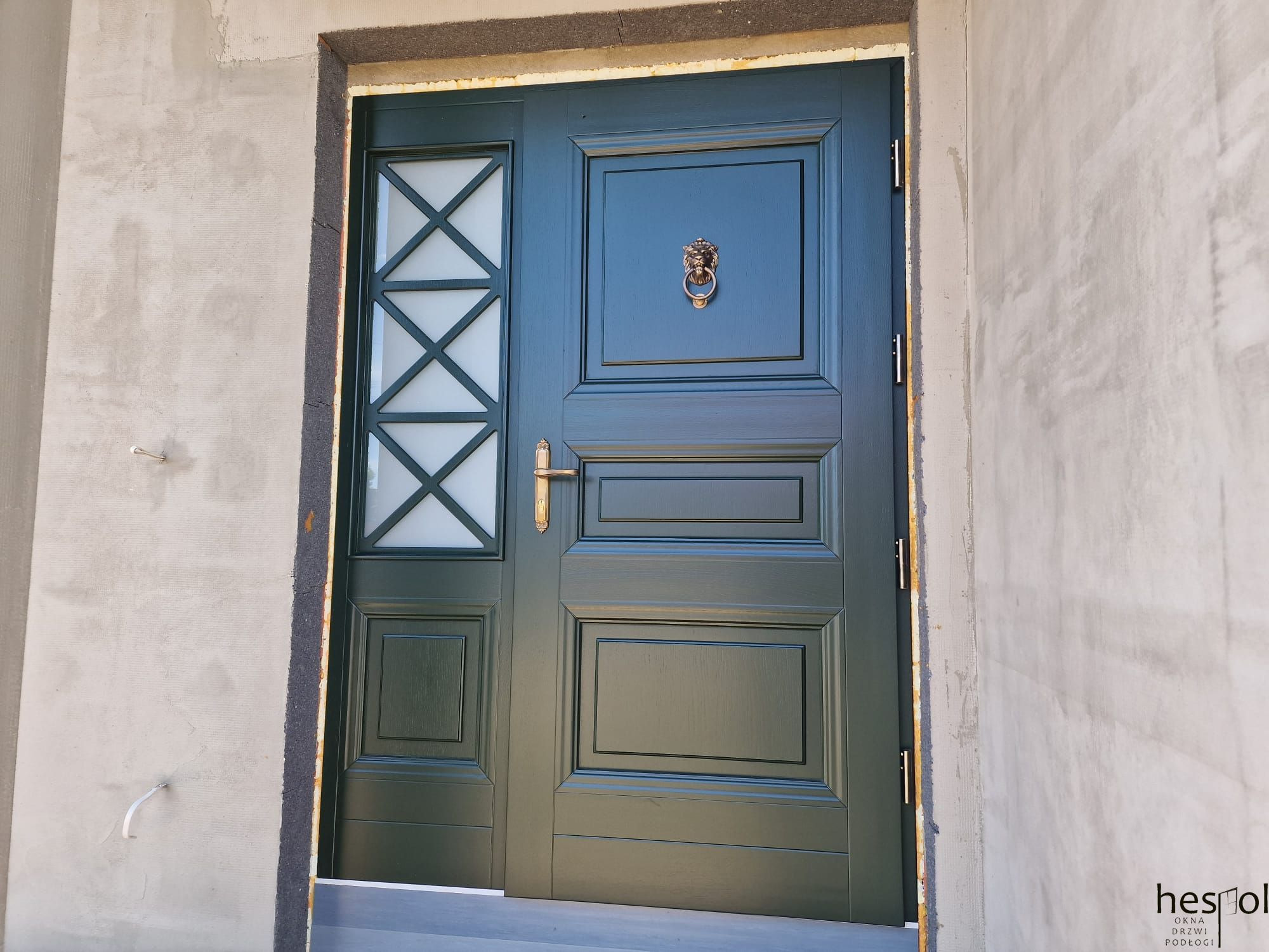 Eleganckie, zielone drzwi zewnętrzne z dekoracyjnym lwem na tle surowego muru. Drzwi posiadają szybę z geometrycznym wzorem i złotą klamkę.