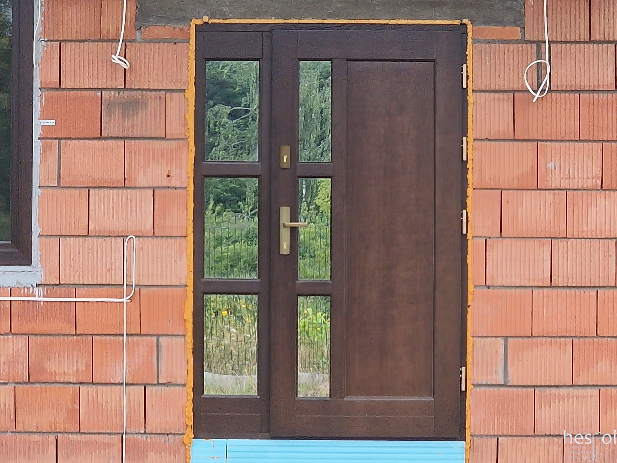 Brązowe drzwi zewnętrzne z szybami zamontowane w surowym murze z cegły, uszczelnione pianką montażową. Widoczny fragment okna i wystające przewody.