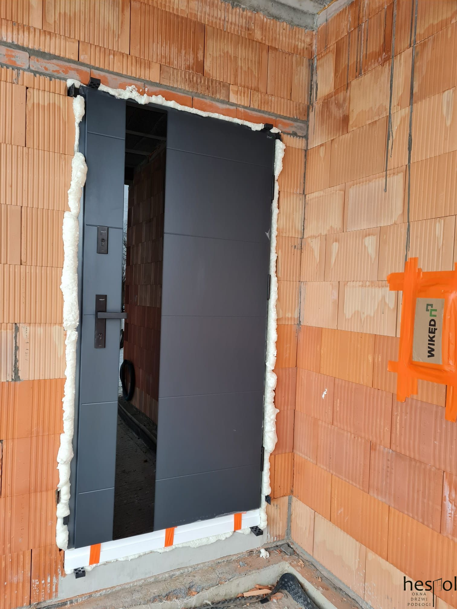 Montaż grafitowych drzwi zewnętrznych w surowym murze z cegły, uszczelnionych pianką montażową. Widoczny fragment wnętrza i detale mocowania.