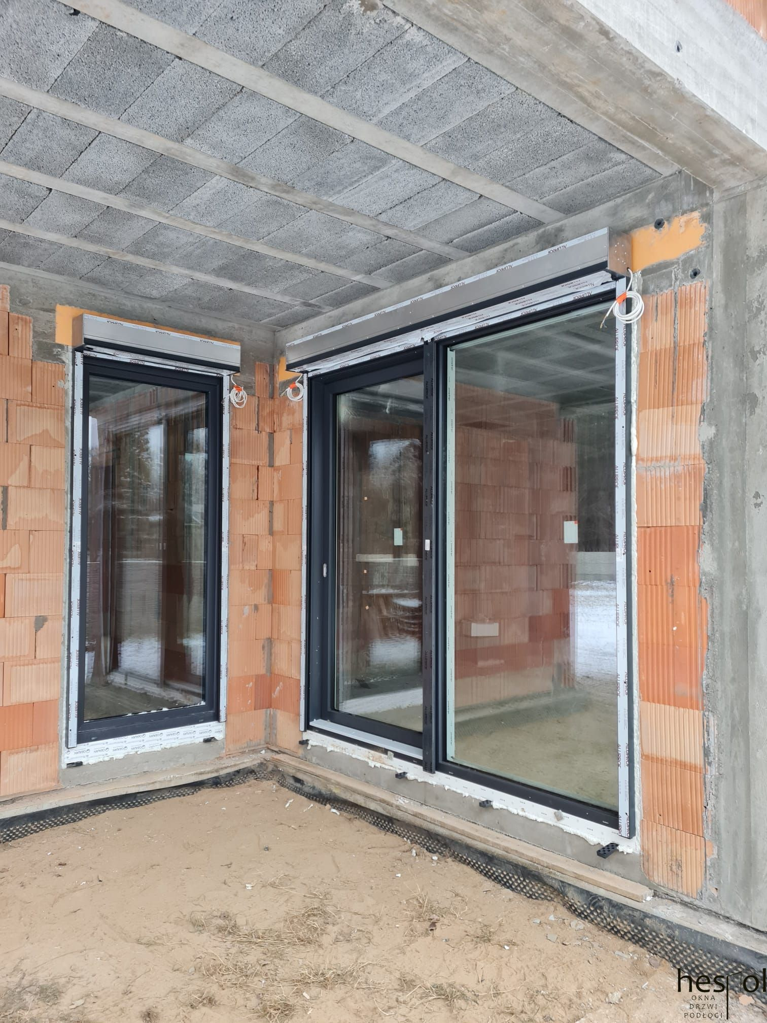 Montaż okien PCV z roletami zewnętrznymi w surowym budynku. Widoczne bloczki betonowe stropu i cegły. Okna z czarnymi ramami i białą listwą montażową.