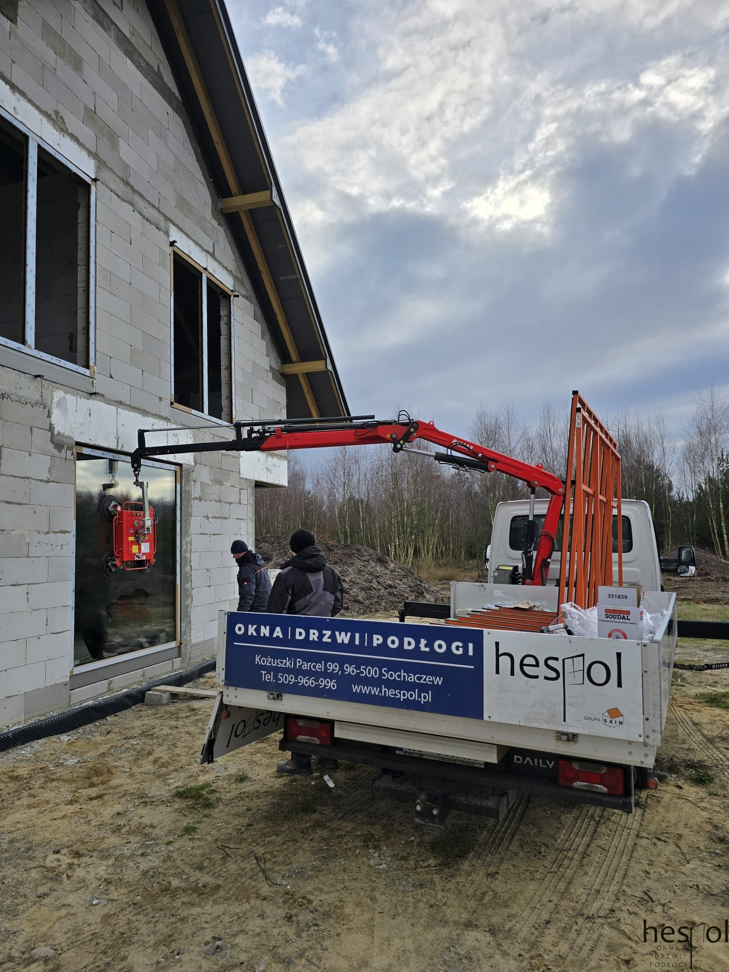 Montaż okna w surowym budynku za pomocą przyssawki i dźwigu, widoczni pracownicy, auto dostawcze z reklamą okien, drzwi i podłóg w tle.