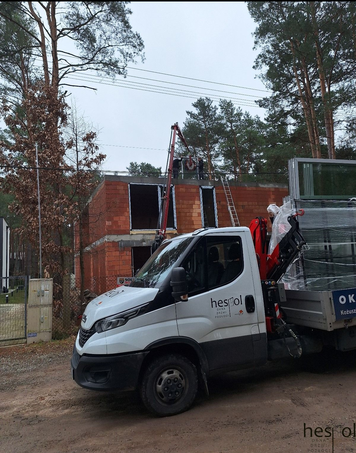 Montaż okien w surowym budynku z użyciem dźwigu. Na pierwszym planie samochód dostawczy firmy z oknami. Widok na plac budowy w pochmurny dzień.