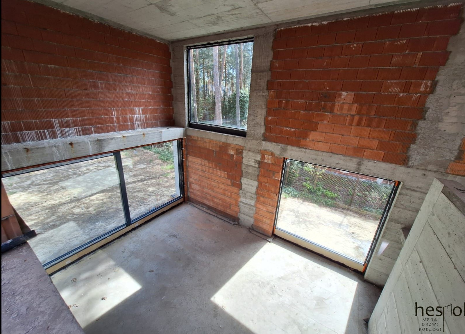Surowy stan: wnętrze z trzema oknami w czarnych ramach, widok na zieleń. Ściany z czerwonej cegły, surowy betonowy strop, wylewka na podłodze. Widok z góry.