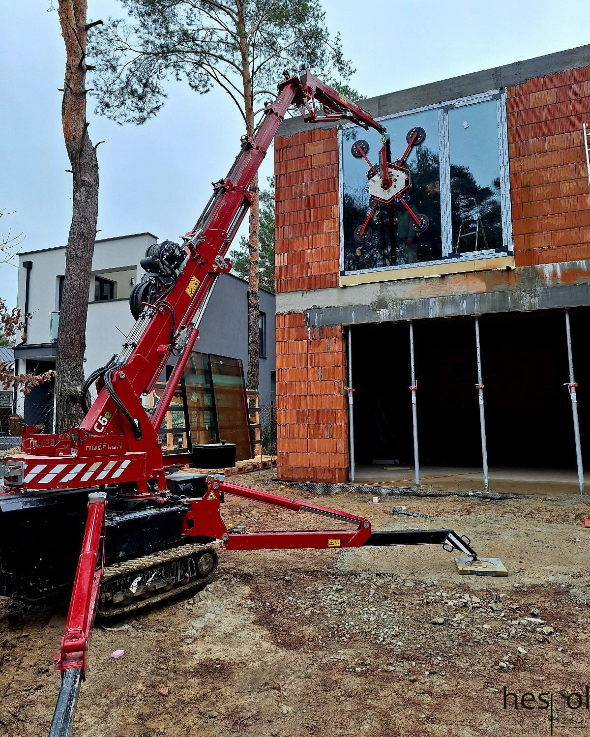 Montaż okna na budowie domu jednorodzinnego za pomocą specjalistycznego dźwigu z przyssawkami. Widoczny surowy stan budynku i otoczenie placu budowy.