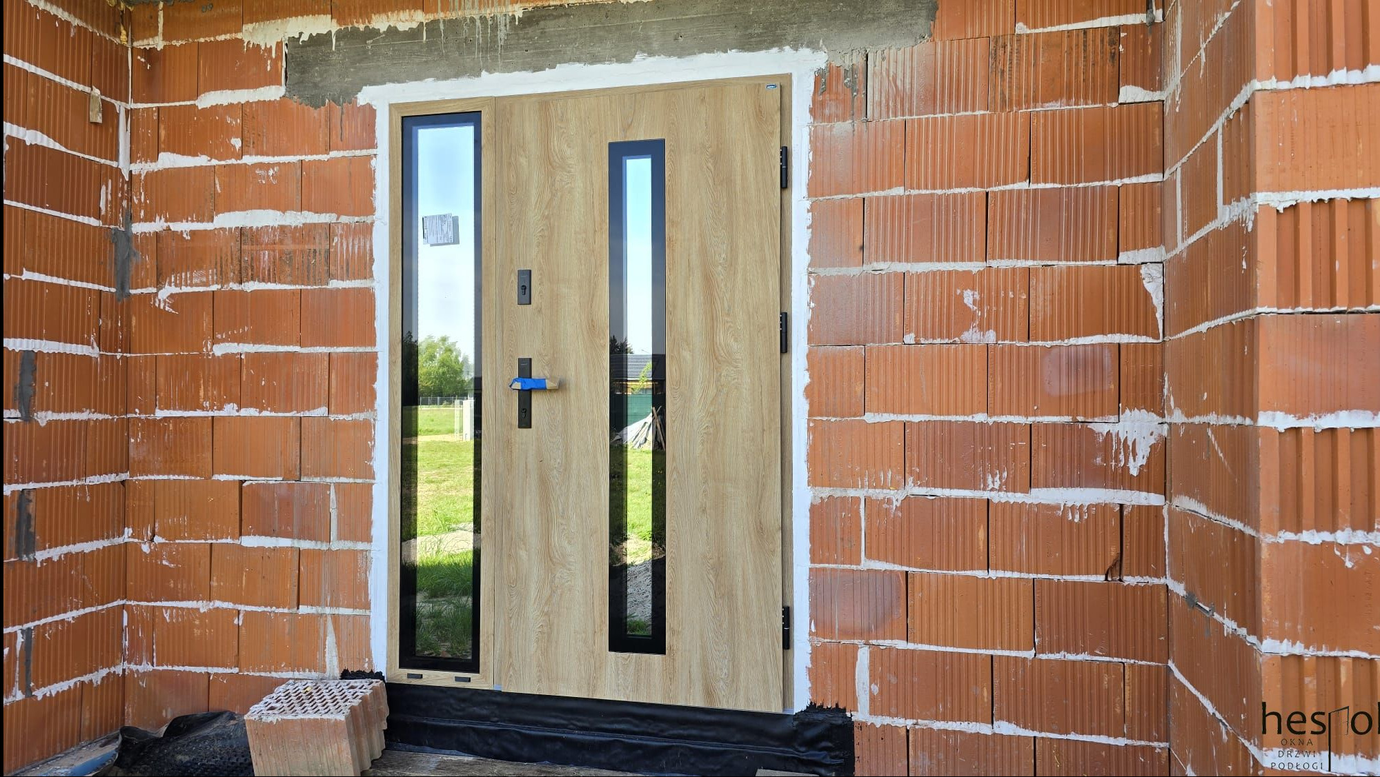 Jasne drzwi zewnętrzne z przeszkleniami, czarnymi ramami i klamką, zamontowane w surowym murze z cegły ceramicznej. Widoczne detale montażu.