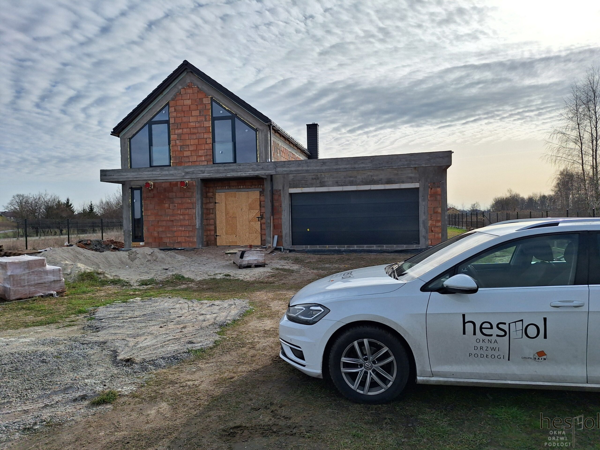 Surowy stan domu z cegły z zamontowanymi oknami, garaż z bramą i auto firmy Hespol na pierwszym planie, pochmurne niebo w tle.