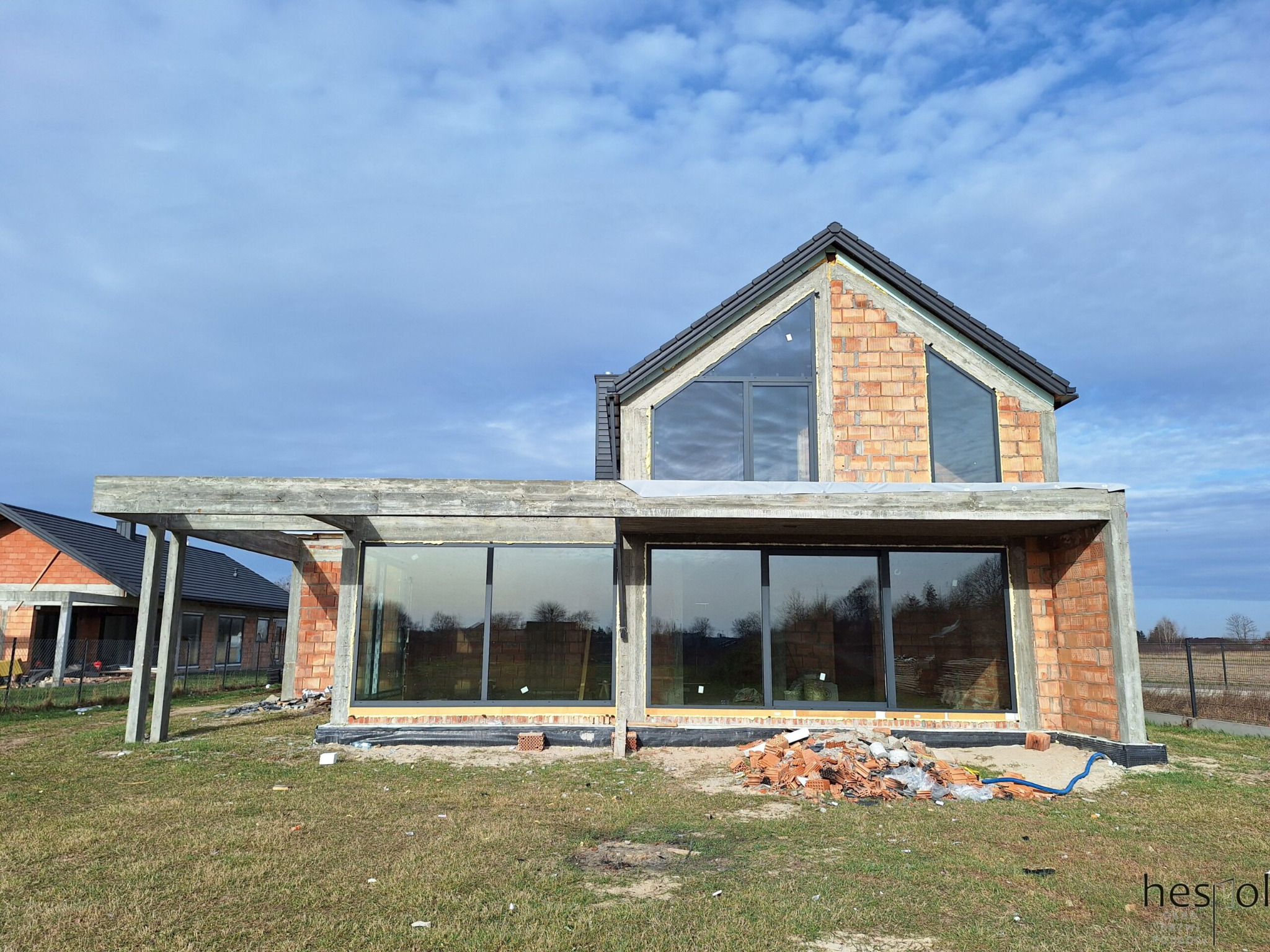 Surowy stan domu z zamontowanymi oknami aluminiowymi, ceglana elewacja w trakcie budowy, widoczne okna z ciemnymi ramami i fragment dachu z szarą dachówką.