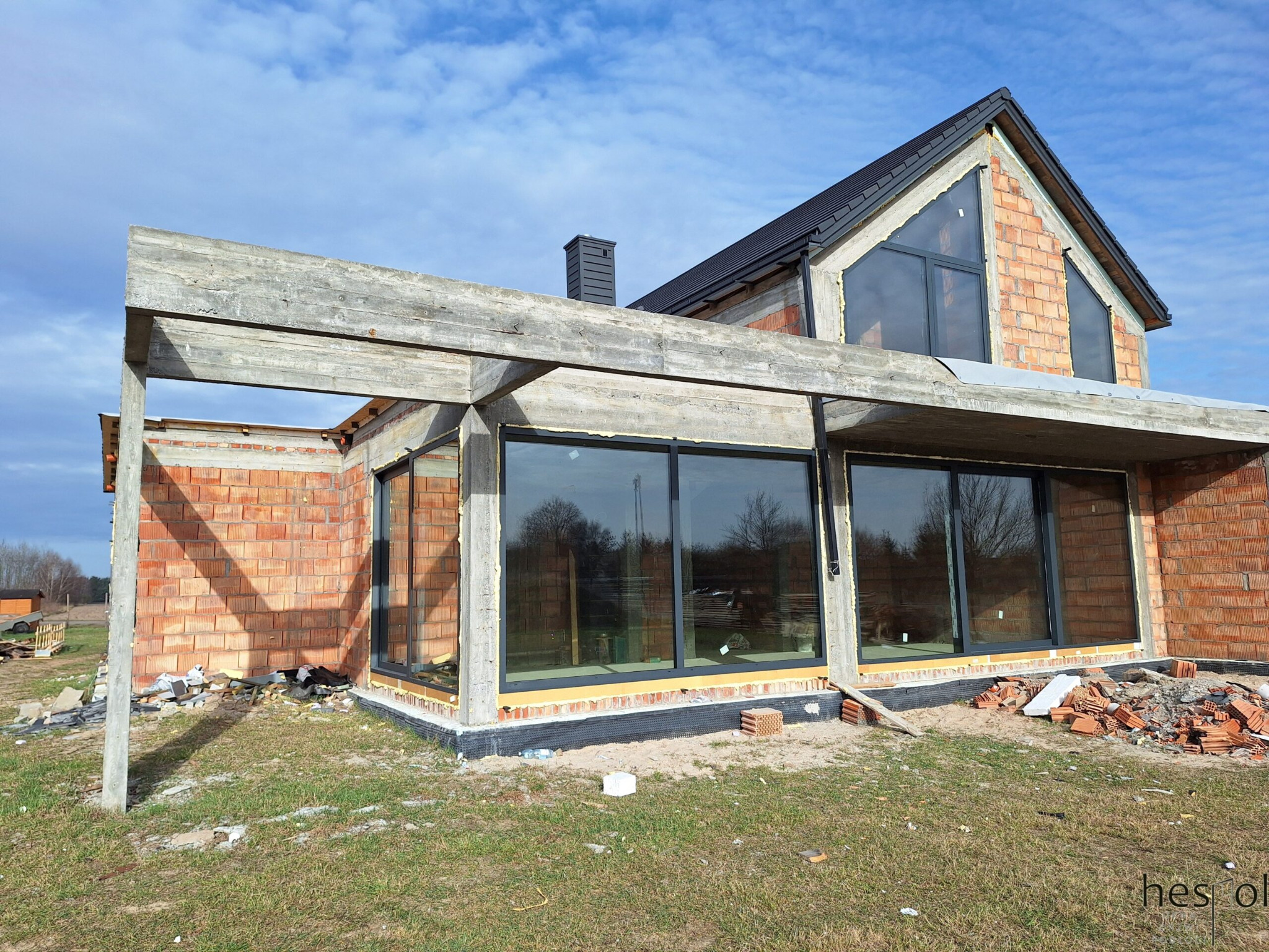 Surowy stan domu z cegły z zamontowanymi oknami z czarnymi ramami, widoczne betonowe elementy konstrukcyjne i fragment dachu, błękitne niebo w tle.