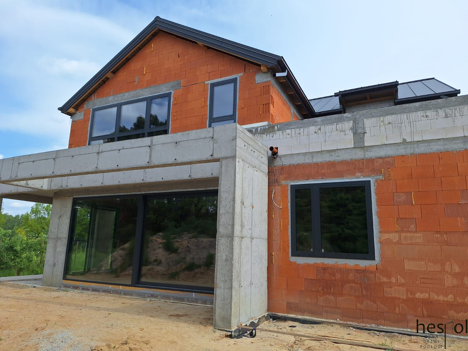 Surowy stan domu z zamontowanymi oknami w Sochaczewie. Widoczne szare ramy okienne w czerwonej, ceglanej ścianie. Duże przeszklenie na parterze.