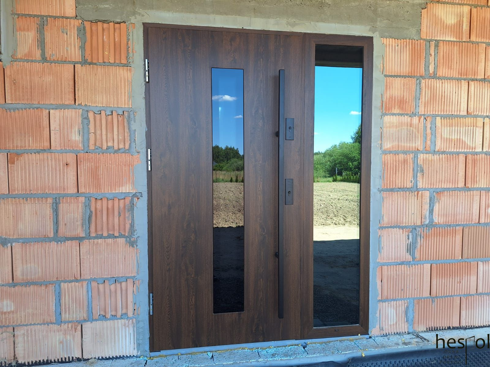 Nowoczesne drzwi zewnętrzne z przeszkleniami w brązowej okleinie, zamontowane w surowym murze z cegły. Długi, czarny uchwyt. Widok na pole i las przez szybę.