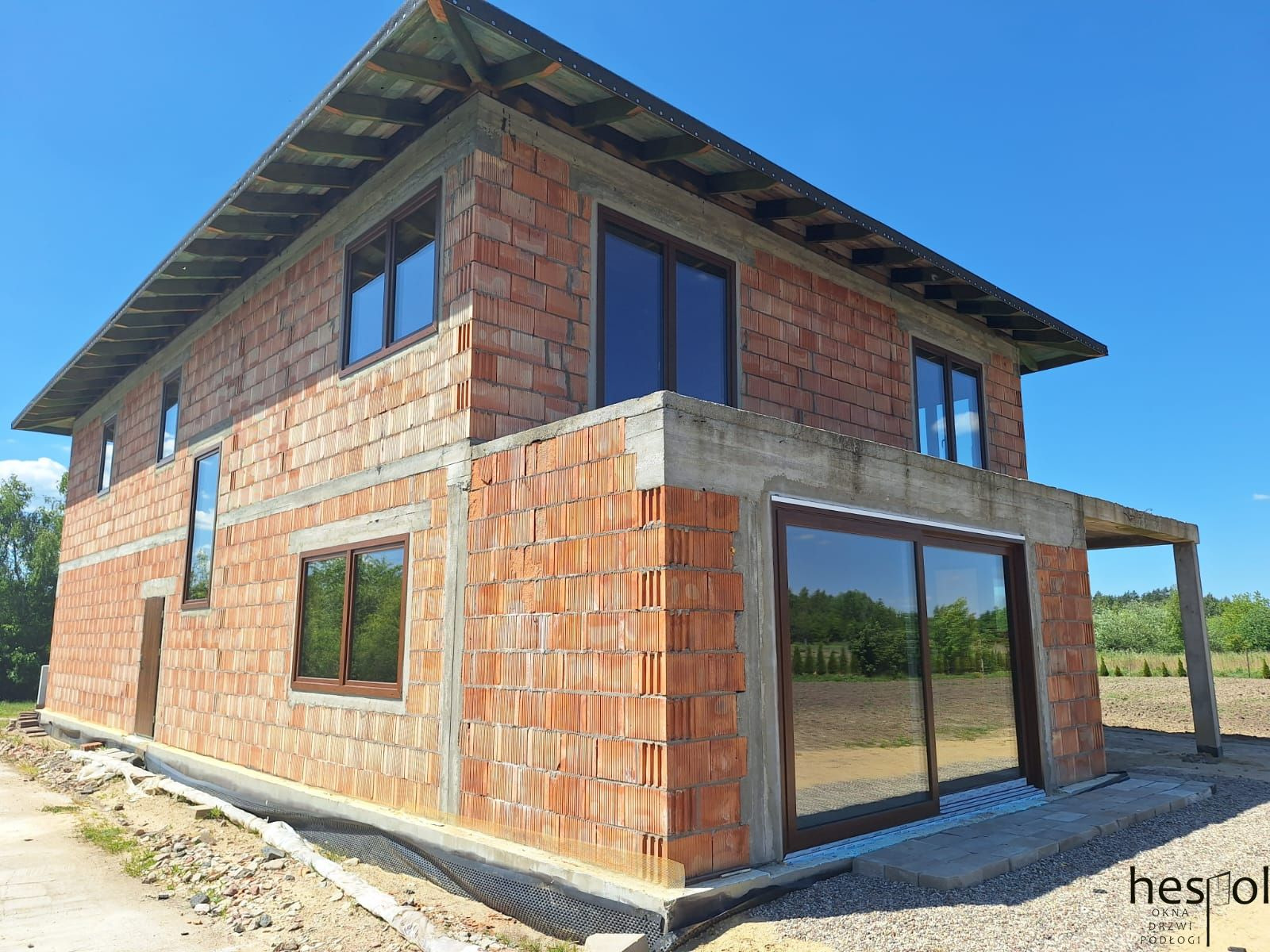 Dom w trakcie budowy z zamontowanymi oknami i drzwiami przesuwnymi w brązowych ramach, widoczne bloczki z czerwonej cegły i szary beton, błękitne niebo.