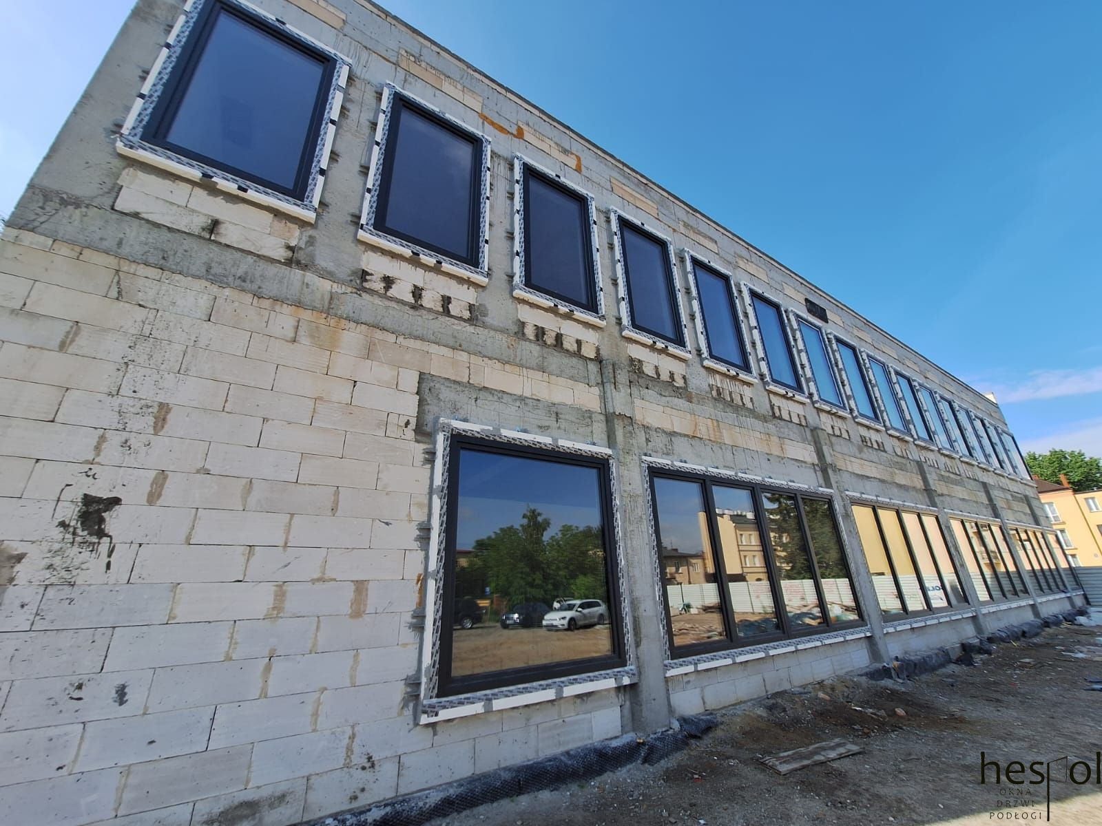 Montaż okien z czarnymi ramami w surowym budynku z betonu komórkowego. Widoczne prace wykończeniowe wokół okien. W szybach odbija się otoczenie.