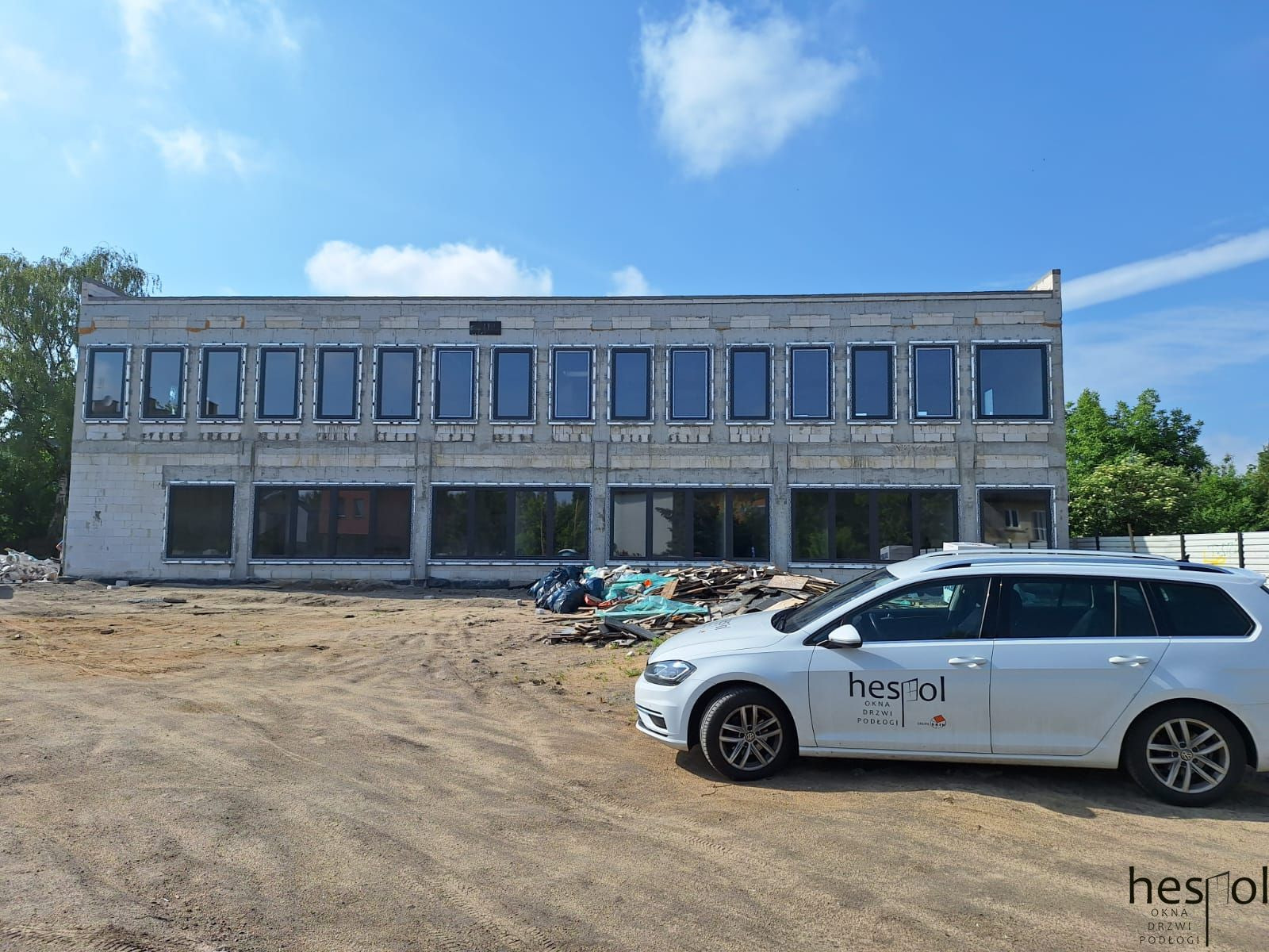 Budynek w trakcie montażu okien z ciemnymi ramami, widoczny samochód z logo firmy. Tło: niebo, drzewa, plac budowy. Ujęcie z boku, w słoneczny dzień.